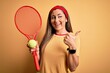 © Krakenimages.com - Beautiful brunette sportswoman playing tennis using racket and ball over yellow background pointing and showing with thumb up to the side with happy face smiling