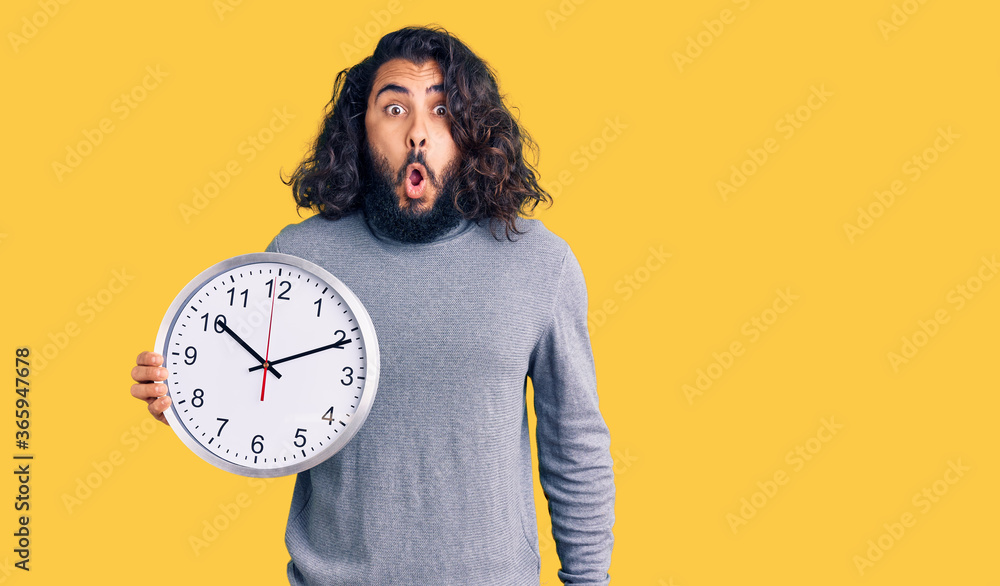 Young arab man holding big clock scared and amazed with open mouth for surprise, disbelief face