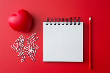 Wall Mural - Top view of Notebook, pencil, paper clip, heart shaped ball  on a red background and copy space