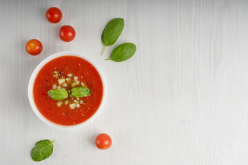 Wall Mural - White bowl full of red gaspacho soup surrounded by whole tomatoes and green basil leaves standing on white wooden table. Image with copy space, horizontal orientation