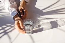 Woman's Hands And A Glass Of Clean Water On The White Table In Natural Sunlight With Plant Shadows.