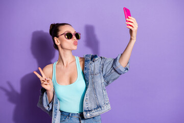 Portrait of charming girl blogger use smartphone make selfie v-sign send air kiss followers subscribers wear denim jeans jacket singlet isolated over violet color background