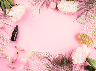 Pink background with beauty lifting rose quartz face roller, essential oil bottle and dry massage brush with floral decor. Flat lay frame