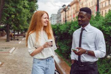 black man and red haired woman entrepreneurs walking outdoors on street and drinking coffee during b