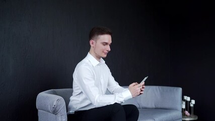  A man sits on a sofa and holds a phone. Writes messages and reads. Smiling