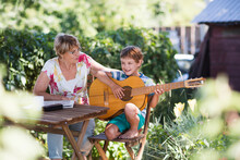 Child Boy Playing  Guitar And Sings Songs With His Grandmother In The Garden.  Elderly Woman With Child Have Fun Together.  Relax Family Activity
