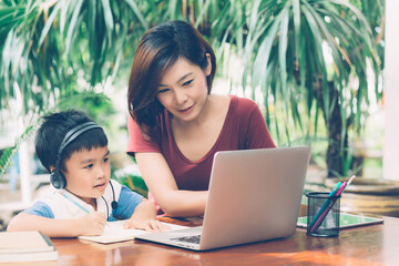 Wall Mural - Young asian mother and son using laptop computer for study and learning together at home, boy writing on notebook for homework and wearing headphone, teacher or mom support child, education concept.