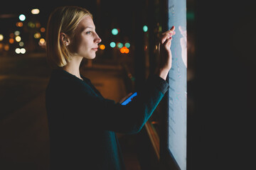 Cropped image young woman touching big digital display while standing in night city with reflected on her screen light, attractive female doing internet money payment with automated teller machine