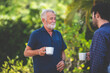 © chokniti - An adult hipster son and senior father drinking coffee in the garden at home, happy talking in the morning