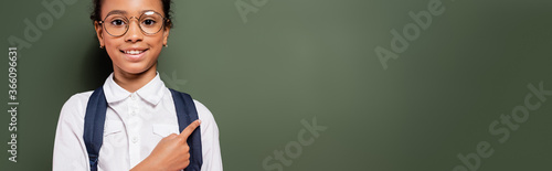 smiling african american schoolgirl pointing at empty green chalkboard, panoramic shot