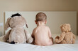 © Cavan Images - Rear view of a naked baby sitting up in bed with two teddy bears