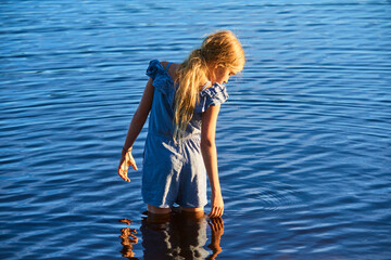 Wall Mural - Alone child blond girl standing in the water in the light of the setting sun in summer