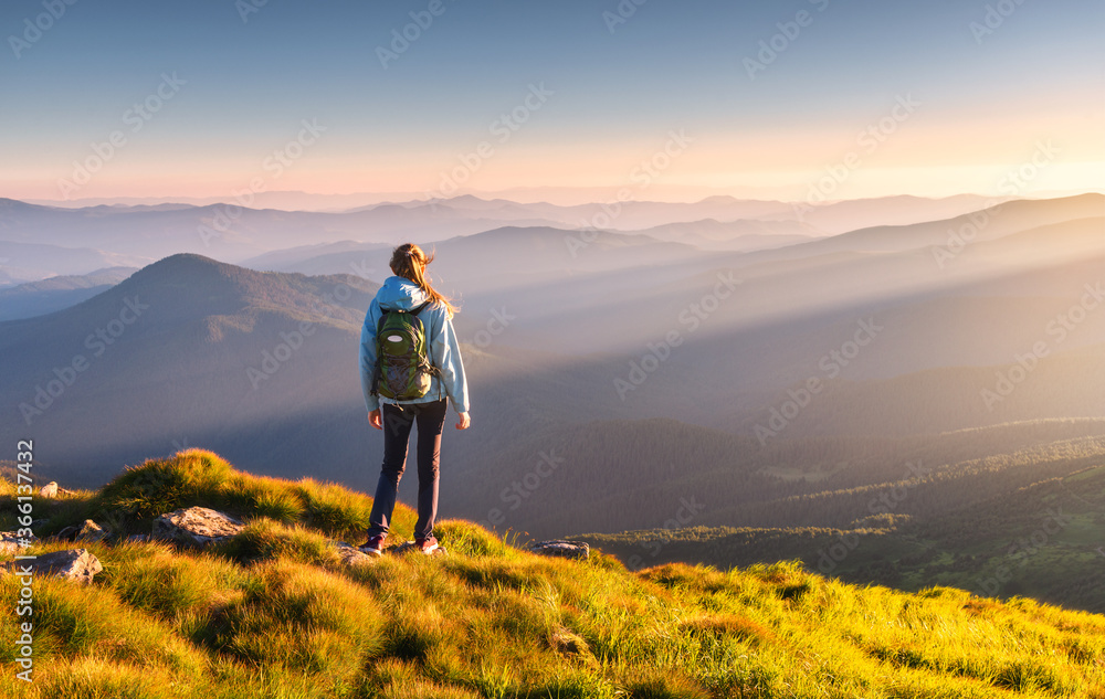 Beautiful mountains in fog and standing young woman with backpack on the peak at sunset in summer. Landscape with sporty girl, green grass, forest, hills , blue sky with sunbeams. Travel and tourism - obrazy, fototapety, plakaty 