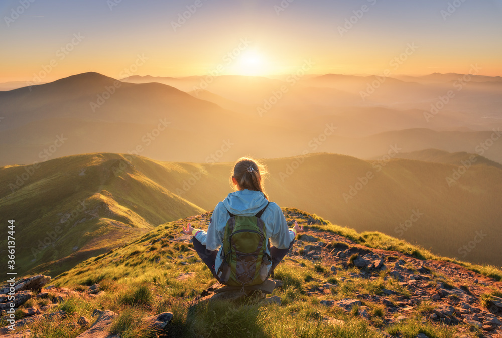 Young woman with backpack sitting on the mountain peak and beautiful mountains in fog at sunset in summer. Landscape with sporty girl, green forest, hills , sky, sunbeams. Travel and tourism. Yoga - obrazy, fototapety, plakaty 