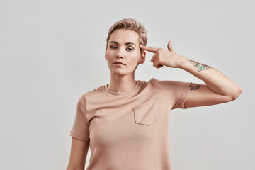 Portrait of tattooed woman with pierced nose and short hair in beige t shirt making suicide gesture, shooting in temple with finger like a gun isolated over light background