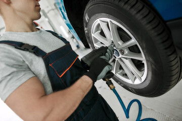 Cropped shot of male mechanic screwing or unscrewing car wheel of lifted automobile by pneumatic wrench at auto repair shop