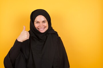 Wall Mural - Beautiful middle aged muslim woman wearing black hijab over yellow background doing happy thumbs up gesture with hand. Approving expression looking at the camera showing success.