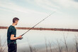 © Westend61 - Side view of teenage boy fishing with rod in lake while standing against cloudy sky