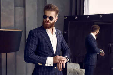 Handsome stylish man in sunglasses and blue suit in a cage at home standing on sofa near mirror. Checking time.