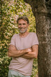 © Westend61 - Smiling bearded mature man standing with arms crossed leaning on tree trunk at garden