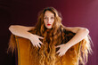 © Westend61 - Portrait of confident young woman with long brown wavy hair leaning on chair against colored background