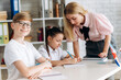 © Kateryna - Lesson at the school. Two schoolchildren, a boy and a girl, are sitting at a desk during a lesson, and a young woman teacher helps them complete the task, smart boy looks at the camera