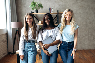 Attractive young girls. African American and Caucasian girls. Beautiful cheerful female classmates stand in a college in stylish clothes looking at the camera and smiling