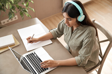 Indian girl student wear headphones learning online watching webinar class looking at laptop computer elearning remote lesson making notes or video calling virtual conference meeting teacher at home.