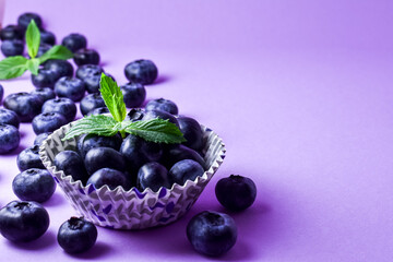 Blueberries in a paper baking dish, two mint leaves on a purple background. Copy space. 