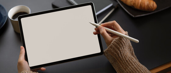 Female hands using mock up tablet with stylus pen on breakfast table with schedule books, coffee and croissant