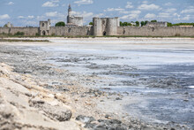 The Ramparts Of The Medieval French Town Of Aigues Mortes Facing Its Salt Pan, Against A Blue Summer Sky With Puffy Clouds