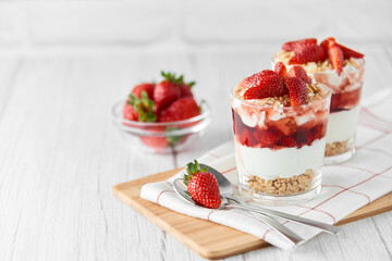 Homemade layered dessert with fresh strawberries, cream cheese or yogurt, granola and strawberry jam in glasses on white wood background. Healthy organic breakfast or snack concept. Selective focus.