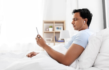 Sticker - technology, bedtime and rest concept - indian man in earphones with smartphone lying in bed and listening to music at home