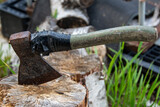 Fototapeta Na sufit - An old rusty ax with a wooden handle is rewound with a black ribbon, stuck in a birch stump against a background of green grass and birch firewood. Chopping.