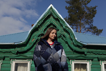 Bohemian, boho girl in vintage patchwork coat. Old wooden rural house with ornamental carved windows, frames in Sergiyev Posad city, Moscow region, Russia. Russian folk style in architecture, fashion