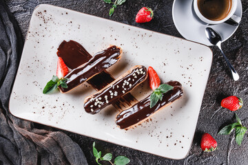 Wall Mural - Homemade cake eclairs or profiteroles with custard, chocolate and strawberries on dark background served with cup of coffee. Traditional french eclairs, dessert. Top view, flat lay