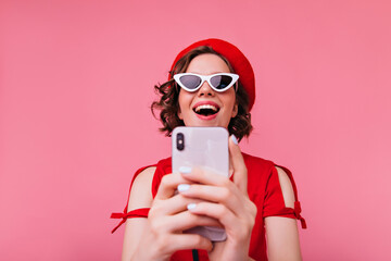 Wall Mural - Funny caucasian girl in french outfit using phone for selfie. Laughing brunette lady in red beret taking picture of herself.