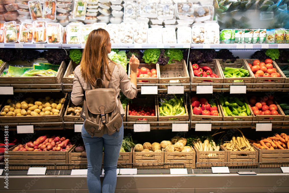 Good looking woman standing in front of vegetable shelves choosing what to buy. Buying groceries and healthy organic food in supermarket. - obrazy, fototapety, plakaty 