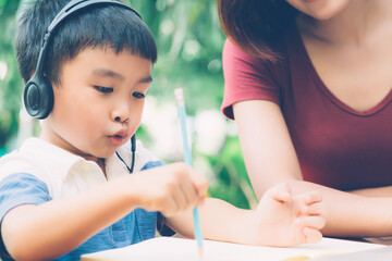 Wall Mural - Young asian mother and son using laptop computer for study and learning together at home, boy writing on notebook for homework and wearing headphone, teacher or mom support child, education concept.