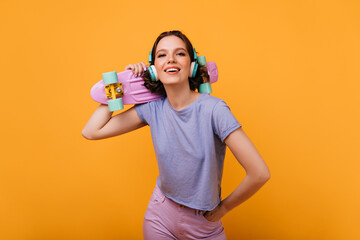 Confident female skateboarder looking to camera with interested smile. Studio shot of pleasant brown-haired woman in headphones isolated on yellow background.