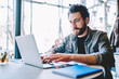 © BullRun - Concentrated bearded young man searching information on website and working on freelance on development graphic project at modern laptop using wireless internet connection in cafe interior