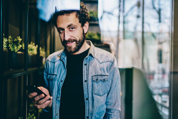 Wall Mural - Portrait of handsome bearded blogger holding smartphone in hand while updating app on device using 4G internet in coworking.Attractive young man chatting on cellular while smiling at camera