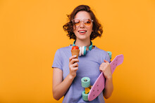 Portrait of smiling white girl holding longboard and eating dessert. Studio shot of brunette glamorous woman with skateboard and ice cream.