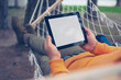 Man lies and rests in a hammock with a mock-up of a tablet with a white screen in his hands.