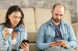© DC Studio - Young husband and wife relaxing in living room using smartphones.