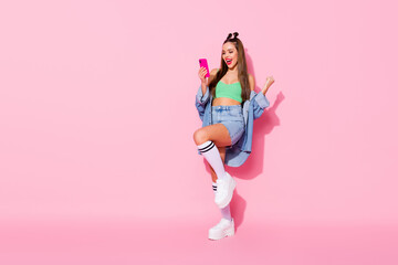 Sticker - Full size photo of pretty excited lady hold telephone read great news raise fist wear denim jacket green crop top mini shirt long socks shoes isolated pastel pink color background