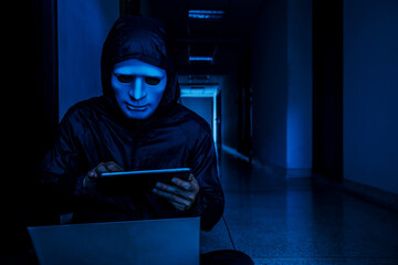 Anonymous computer hacker in white mask and hoodie. Obscured dark face working on her labtop and tablet on target room background, Data thief, internet attack, darknet and cyber security concept..