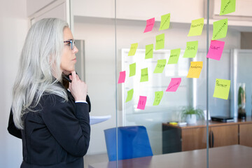 Wall Mural - Confident concentrated businesswoman looking at stickers on glass wall. Focused grey-haired female worker thinking about notes for project strategy. Marketing, business and management concept