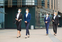 Group Of Female Managers In Office Suits And Masks, Walking Together Past City Building, Talking, Discussing Projects. Full Length Business During Covid Epidemic Concept