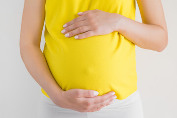 Wall Mural - Young woman in yellow clothes touching big belly with hand. Isolated on gray background. Emotional loving pregnancy time - 33 week. Baby expectation. Happiness and safety concept. Front view. Closeup.